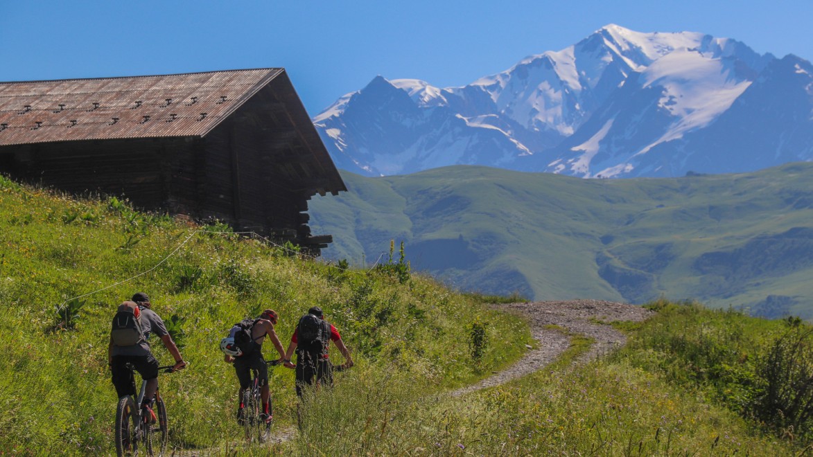 un groupe de VTT passe au pied d'un chalet avec le Mont Blanc en fond