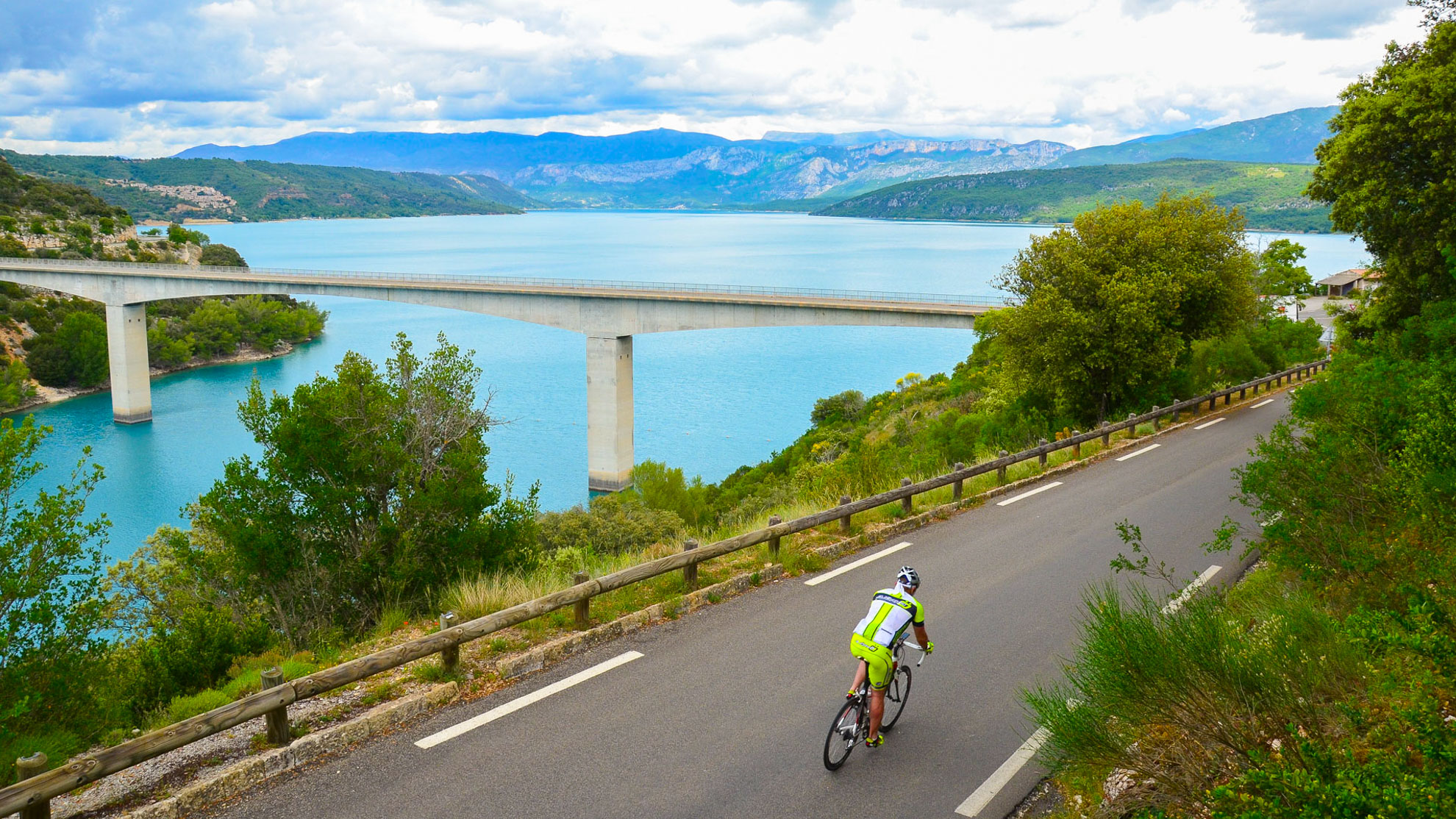 un cycliste sur une route goudronnée longeant le lac de Sainte-Croix, avec le pont sur sa sa gauche