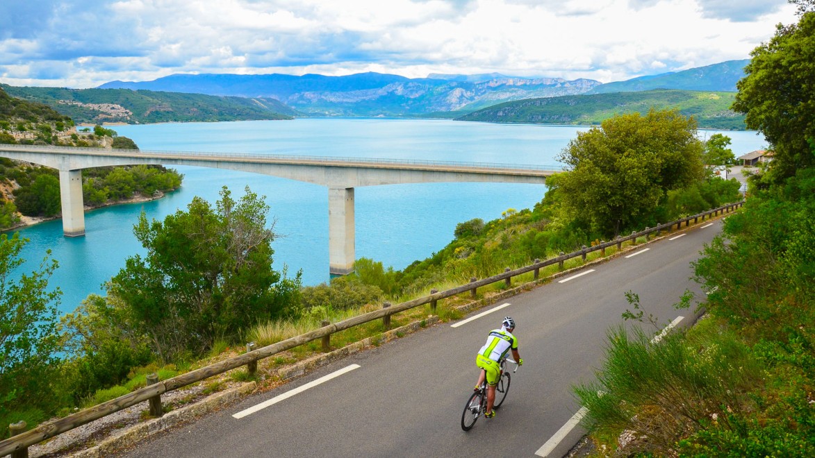 un cycliste sur une route goudronnée longeant le lac de Sainte-Croix, avec le pont sur sa sa gauche