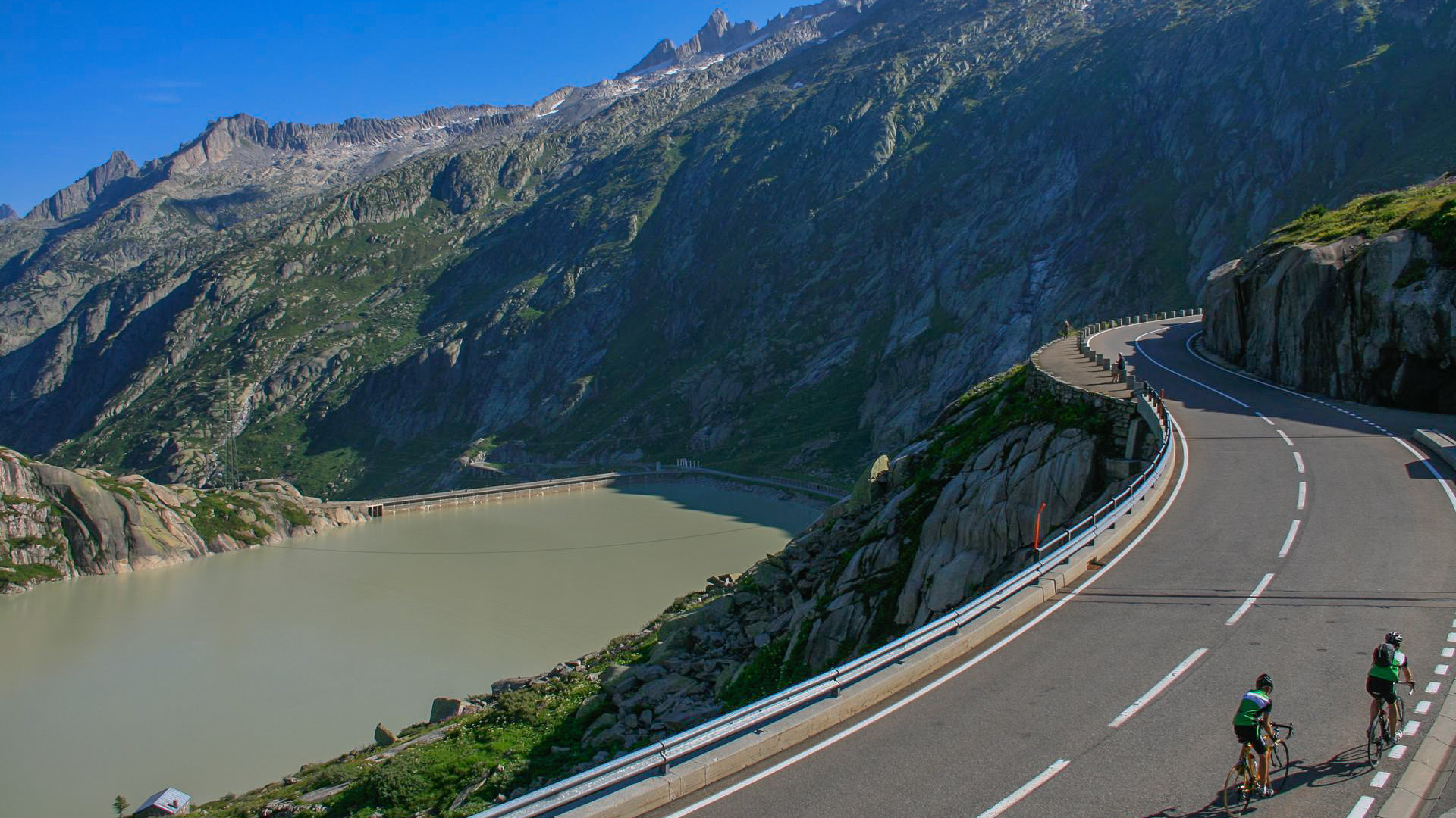 deux cyclistes qui font l'ascension du col de Grimsel en Suisse, avec sur leur gauche le lac de Grimsel