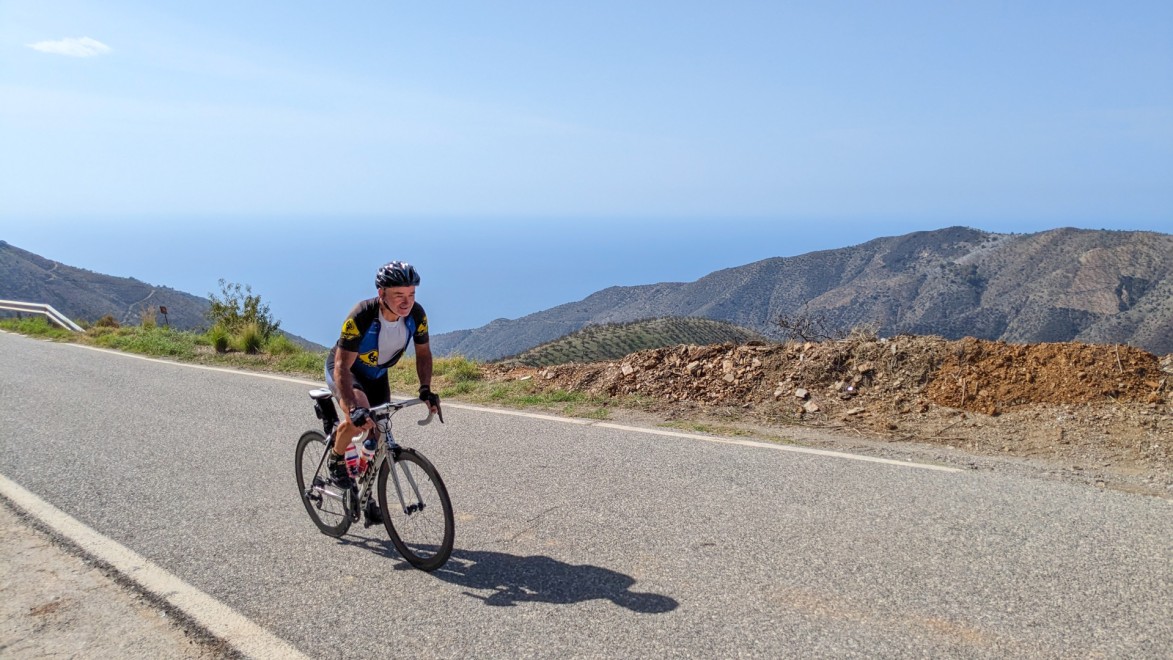 cycliste en danseuse qui monte un col, avec en arrière plan la vue sur la mer Méditerranée