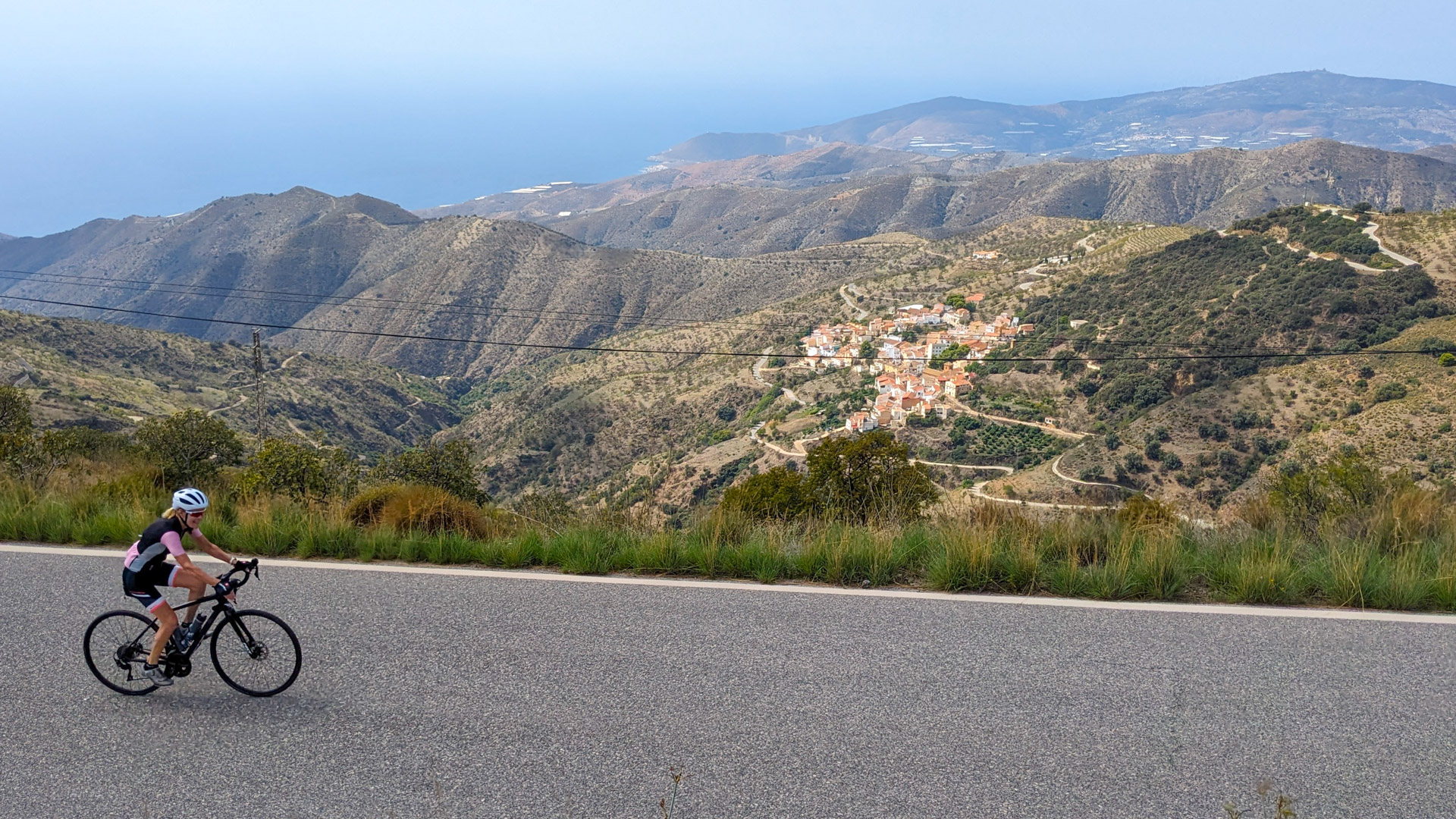 une cycliste sur une route goudronnée, surplombant une petit village de caractère perdu au milieu des montagnes