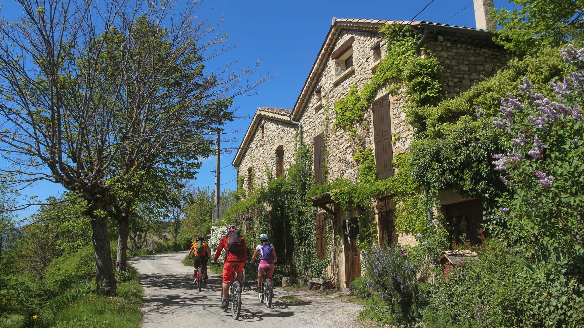 des gens à VTTAE passent devant une maison en pierre typique de la Drôme