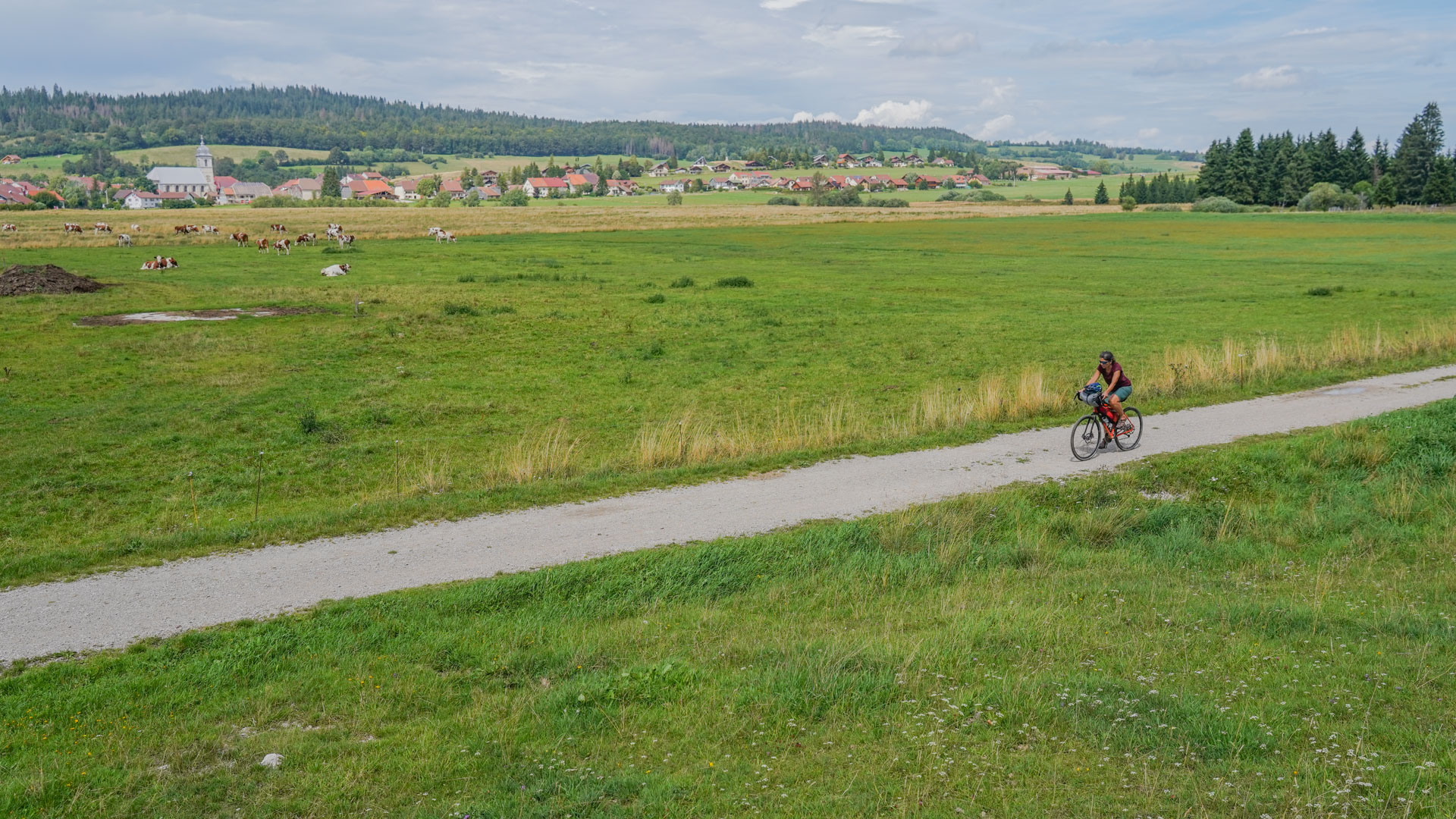 cycliste qui pédale sur une petite route dans la plaine avec un village jurassien en fond