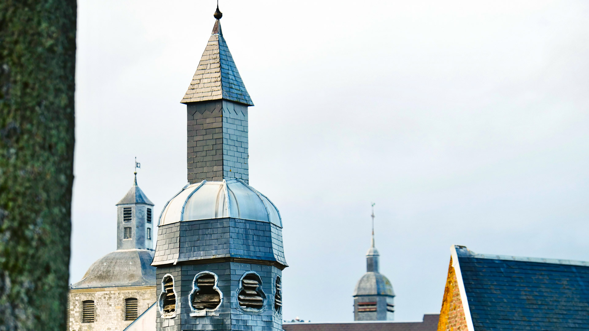 3 toits de ce qui semble être une chapelle, ou un château, sur la droite, un toit plus traditionnel est visible, il est de couleur orange; et contraste avec les autres toits.