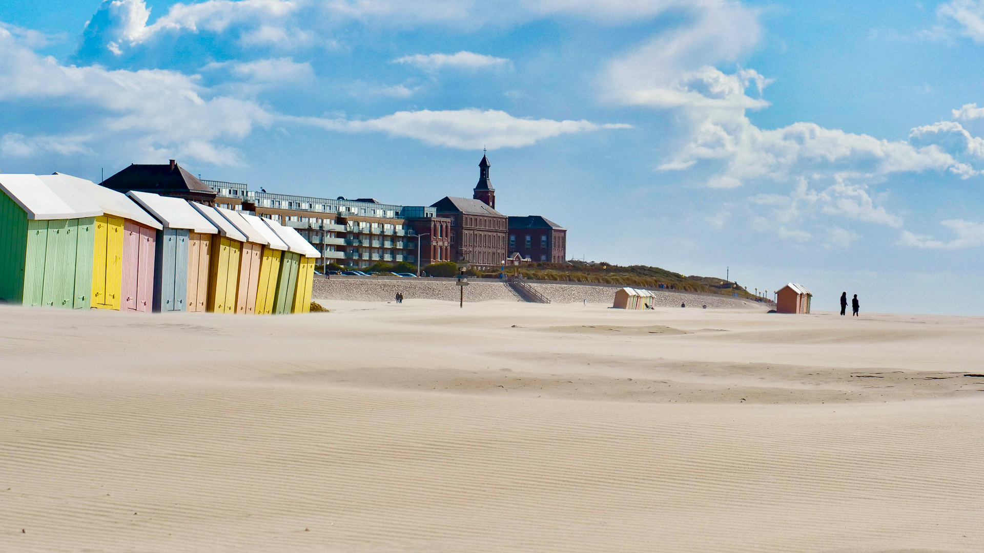la plage s'étend sur tout le bas de l'image, et sur la gauche, nous pouvons apercevoir plusieurs cabines de couleurs vives. En arrière plan, on voit un château de couleur plus foncée