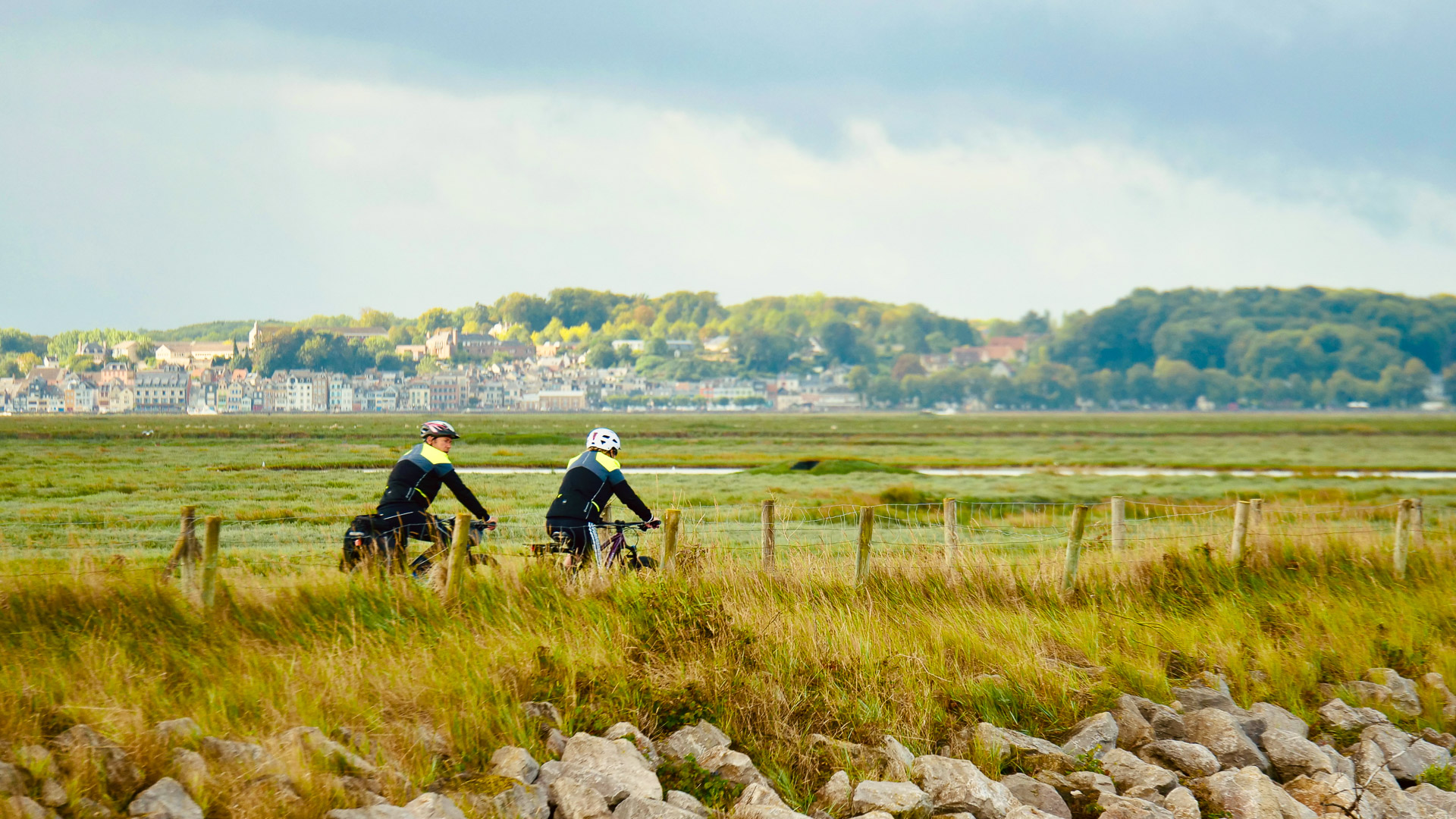 deux personnes à vélo sur une route de campagne, on peut voir au loin des bâtiments de la ville