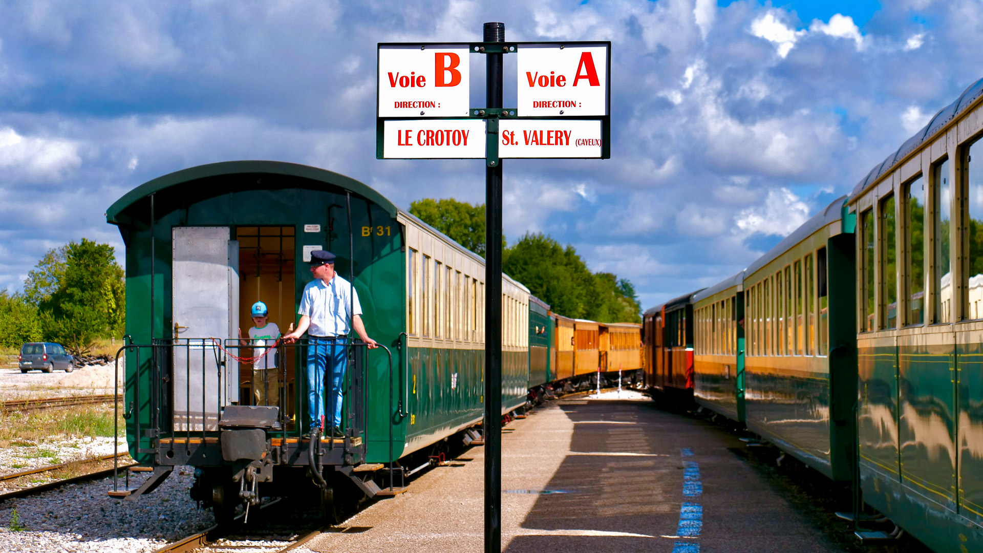 au centre de l'image on voit un panneau indiquant les voies, à gauche on voir une enfant à l'entrée d'un train, accompagnée d'un adulte