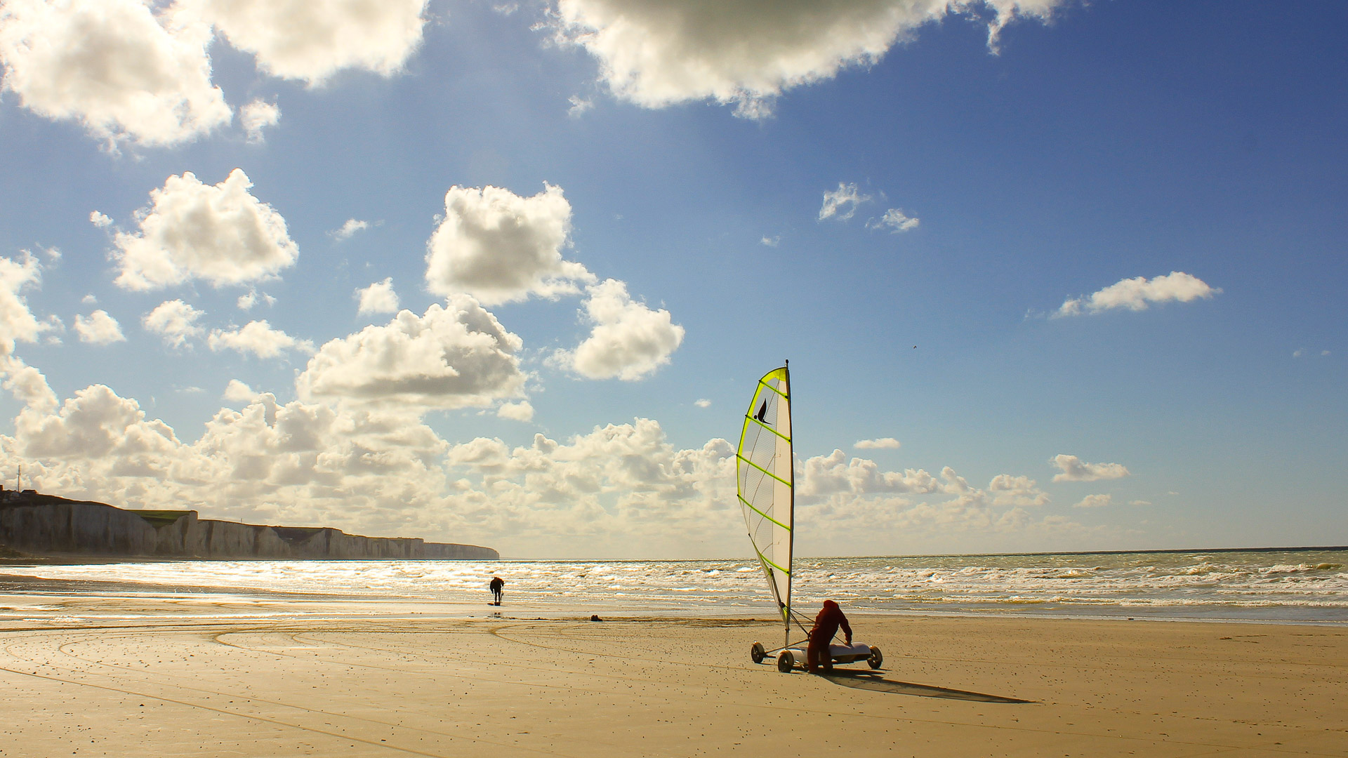 un char à voile sur la plage, un homme grimpe à l'intérieur, à l'arrière plan, on voit des nuages blancs flotter dans le ciel bleu