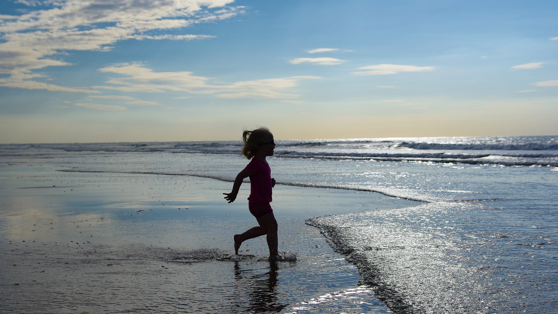 une jeune fille cours vers la mer, le sable est coloré par l'eau, et le ciel bleu derrière la silhouette de la jeune fille souligne la beauté du lieu