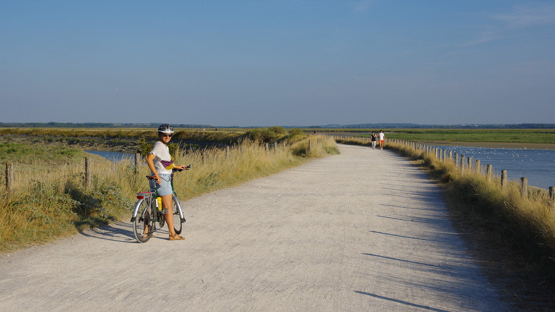 une large route graveleuse entourée de hautes herbes et d'eau, au milieu de laquelle une femme se tient debout sur son vélo, et regarde le photographe