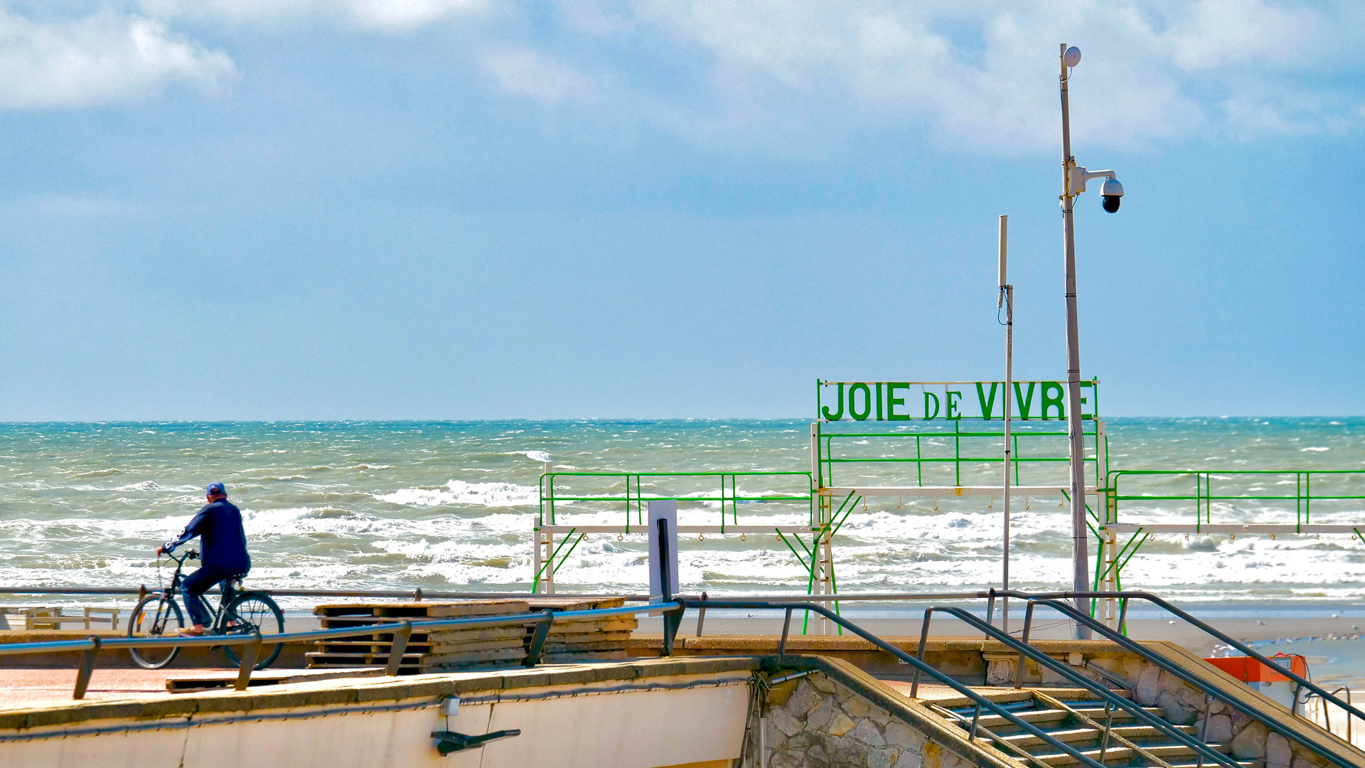 sur la gauche de l'image, on voit un cycliste qui regarde l'horizon. On voit également la mer, et le ciel très bleu; ainsi qu'un panneau 