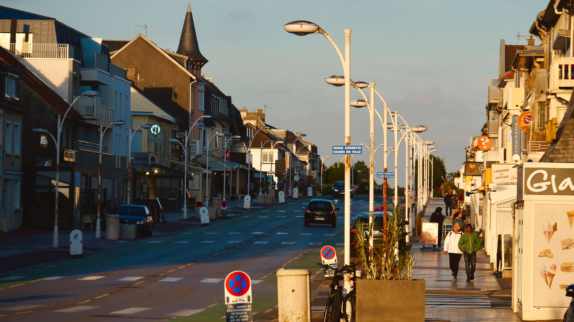 une photo de la ville, avec un bel éclairage. On peut voir des piétons, des voitures, différents commerces, et des lampadaires