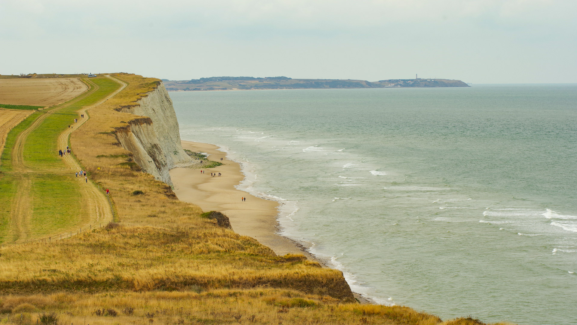 sur la gauche de l'image, on peut voir une grande étendue d'herbe, avec un petit sentier, sur lequel on aperçoit des piétons et des cyclistes. cette étendue s'arrête en allant vers la droite, sur une falaise. En bas de celle-ci, une petite étendue de sable; et la mer