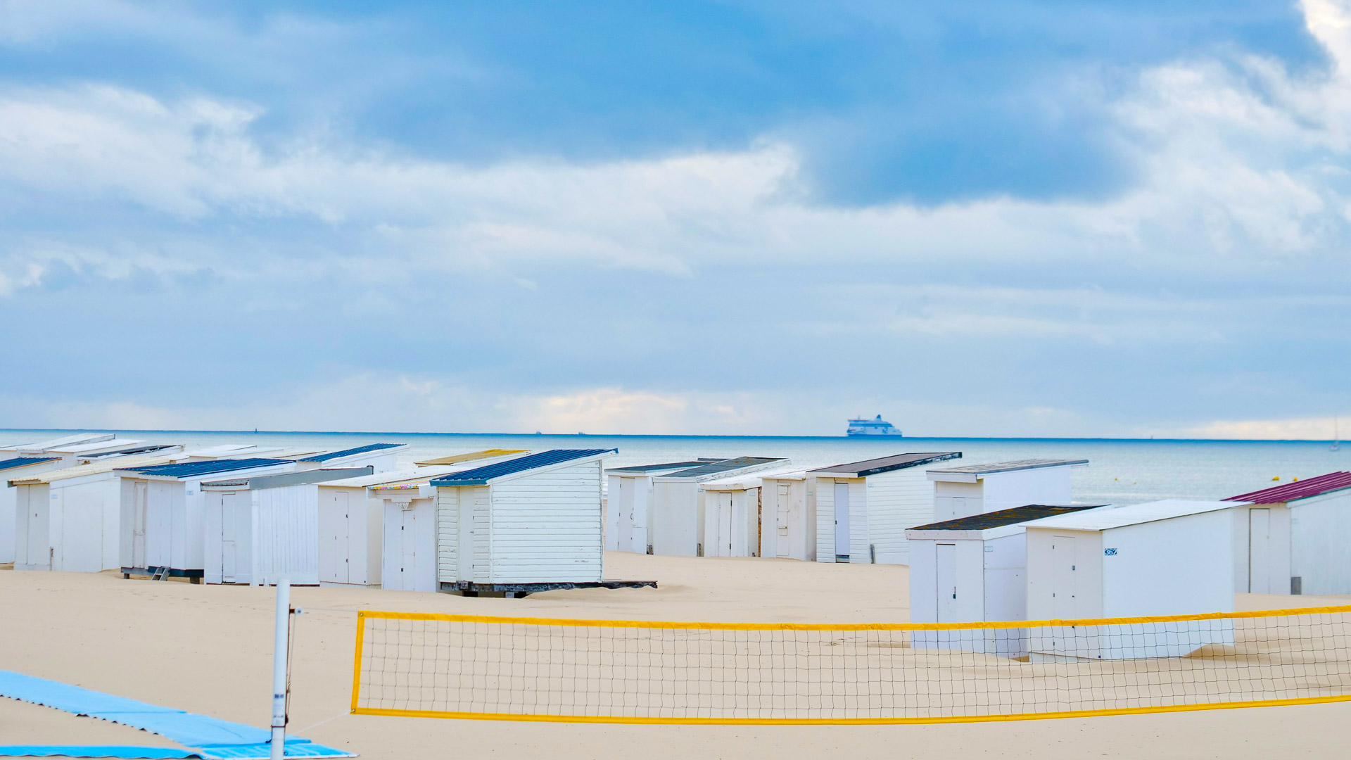 un filet de volley-ball est tendu en bas de l'image. derrière celui-ci, on voit des cabines sur la plage, et au troisième plan, on peut voir la mère, et un navire de croisière