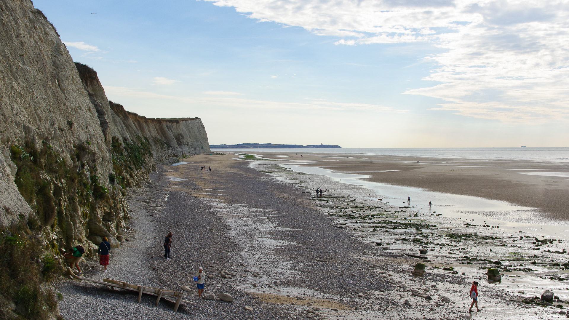 sur la photo, on peut voir la plage, avec différentes personnes dessus. Sur la gauche, on voit une falaise, et sur la droite, on voit la mer