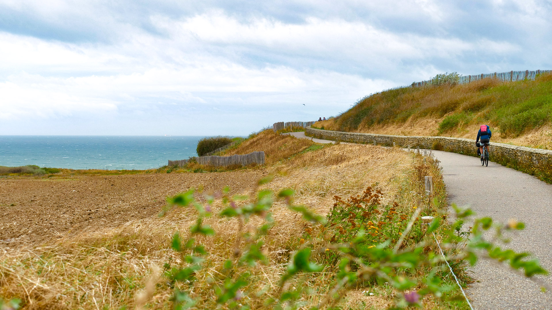 au premier plan, on peut apercevoir des plantes; sur la droite de l'image, on peut voir un cycliste sur une route goudronnée, encadrée par des herbes à gauche, et un muret en pierre à droite. Au fond à gauche de l'image, on aperçoit la mer