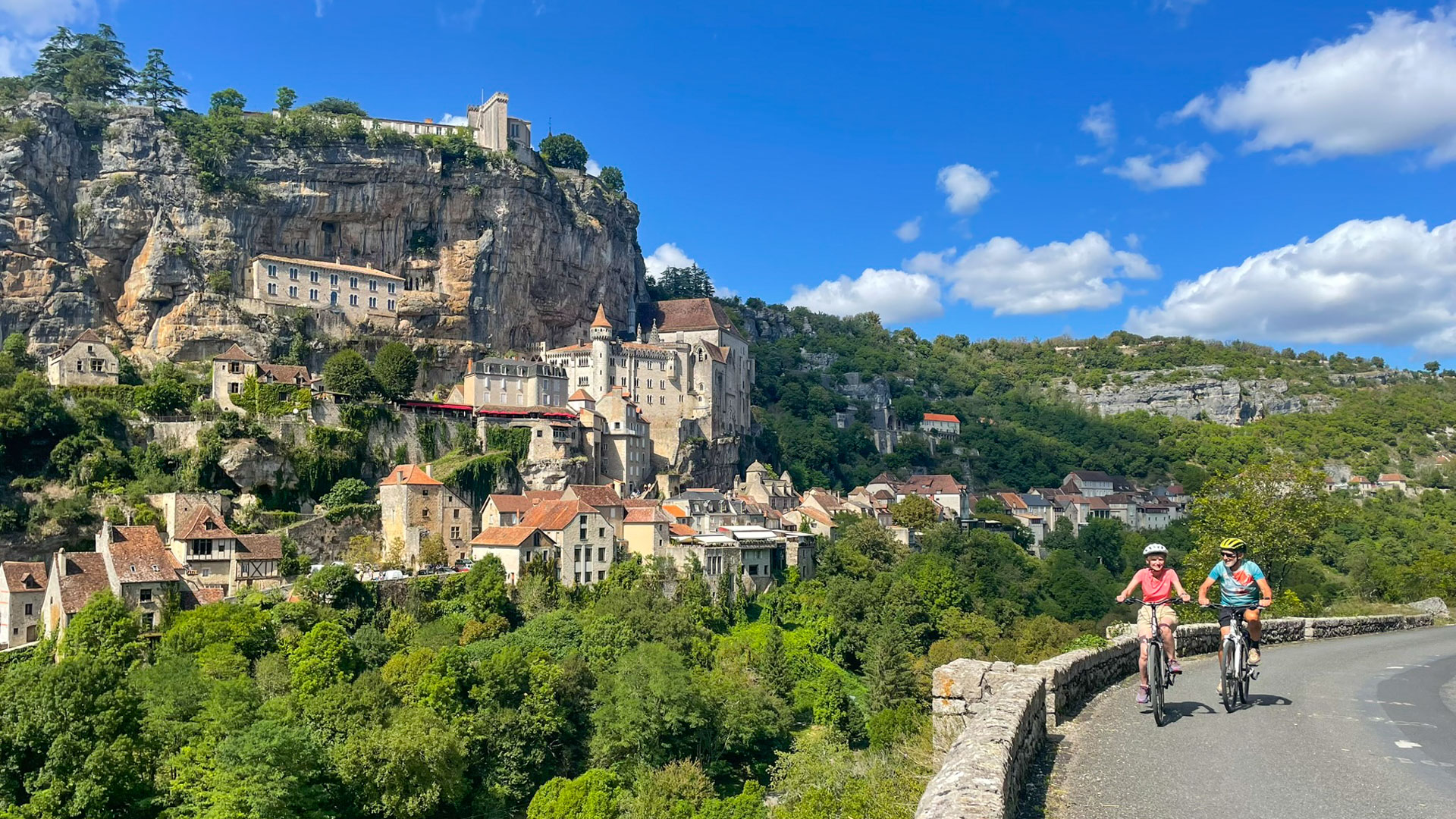 couple de voyageurs à vélo sur une petite route avec en arrière-plan le sanctuaire de Rocamadour